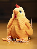 A bright yellow chicken with overlapping fabric feathers, flappy feet and embroidered eyes sits on a wooden surface. A white Sacred Pet Boutique watermark.