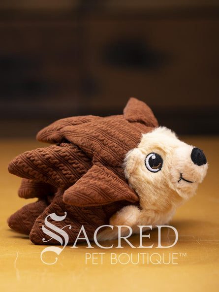 A brown happy hedgehog with overlapping fabric quills, fabric feet and embroidered eyes and mouth sits on a wooden surface. A white Sacred Pet Boutique watermark.