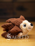A brown happy hedgehog with overlapping fabric quills, fabric feet and embroidered eyes and mouth sits on a wooden surface. A white Sacred Pet Boutique watermark.