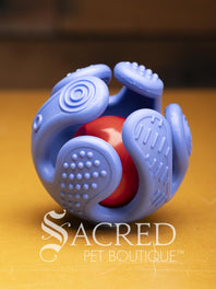 A purple hand shaped ball with textured fingers that curl around a red treat ball sits on a wooden surface. A white Sacred Pet Boutique watermark.