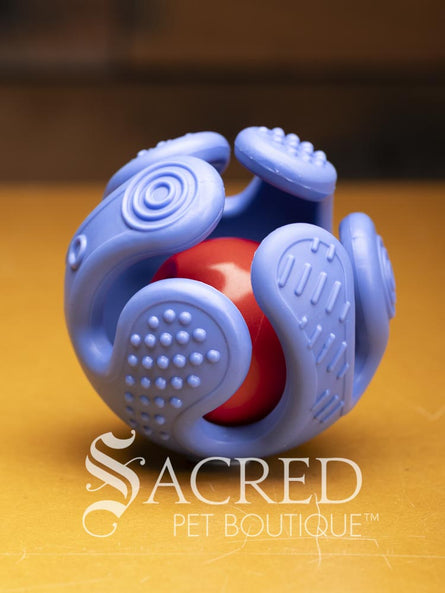 A purple hand shaped ball with textured fingers that curl around a red treat ball sits on a wooden surface. A white Sacred Pet Boutique watermark.