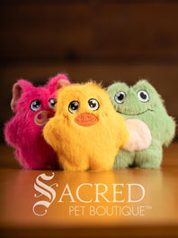 Three colorful plush toys (pink, yellow, green) on a wooden surface with 'Sacred Pet Boutique' branding.