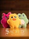 Three colorful plush toys (pink, yellow, green) on a wooden surface with 'Sacred Pet Boutique' branding.