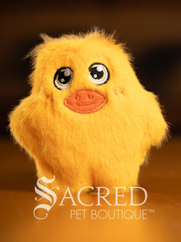 Three colorful plush toys (pink, yellow, green) on a wooden surface with 'Sacred Pet Boutique' branding.