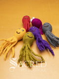 A collection of colourful felt toys in octopus shapes. Included are a yellow, green, purple, grey pink and orange octopus. Displayed on a light wood surface with a white Sacred Pet Boutique watermark. 