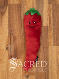 A red furry chilli pepper cat toy with a happy smiling face and a green top or hat sits on a wooden surface.