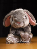 A plush gray rabbit toy with floppy pink inner ears, photographed against a dark background on a wooden surface. A large 'Sacred Pet Boutique' watermark is overlaid across the lower portion of the image.