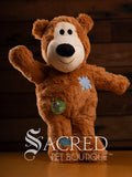 Brown teddy bear with a patch and tag, standing on a wooden surface with a blurred background.