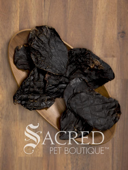 Dark, dried lamb dog treats arranged on a light wood bowl over a wooden tabletop, with the Sacred Pet Boutique logo overlaid in white at the bottom.
