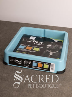 A light blue LickiMat Keeper pet lick mat tray with its retail packaging insert, placed on a gray countertop against a white wall. A watermark for 'Sacred Pet Boutique' is visible at the bottom.