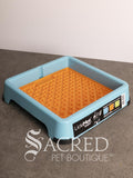 A blue LickiMat keeper with an orange Buddy lickimat tray, shown on a grey surface against a white background. A watermark for 'Sacred Pet Boutique' is visible at the bottom.