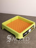A green LickiMat keeper with an orange Buddy lickimat tray, shown on a grey surface against a white background. A watermark for 'Sacred Pet Boutique' is visible at the bottom.