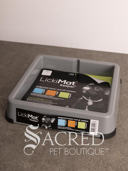 A light blue LickiMat Keeper pet lick mat tray with its retail packaging insert, placed on a gray countertop against a white wall. A watermark for 'Sacred Pet Boutique' is visible at the bottom.