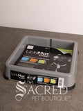 A grey plastic LickiMat Keeper on a grey floor against a white wall, with the product packaging visible inside the mat and a large 'Sacred Pet Boutique' watermark across the lower portion of the photo.