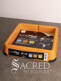 An orange LickiMat Keeper on a grey surface against a white wall, with a product label visible on the front and a large 'SACRED PET BOUTIQUE' watermark at the bottom.