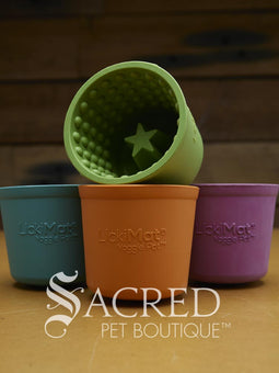 Four LickiMat Yoggie Pots of different colours are lined up on a wooden base with a wooden background. There is a teal, orange and purple one standing upright with a green one lying on top. There is a white Sacred Pet Boutique watermark at the bottom of the image. 