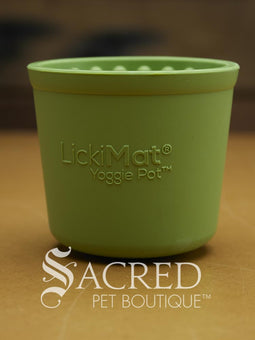 Four LickiMat Yoggie Pots of different colours are lined up on a wooden base with a wooden background. There is a teal, orange and purple one standing upright with a green one lying on top. There is a white Sacred Pet Boutique watermark at the bottom of the image. 