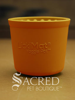 Four LickiMat Yoggie Pots of different colours are lined up on a wooden base with a wooden background. There is a teal, orange and purple one standing upright with a green one lying on top. There is a white Sacred Pet Boutique watermark at the bottom of the image. 