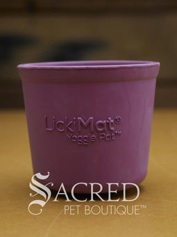 Four LickiMat Yoggie Pots of different colours are lined up on a wooden base with a wooden background. There is a teal, orange and purple one standing upright with a green one lying on top. There is a white Sacred Pet Boutique watermark at the bottom of the image. 