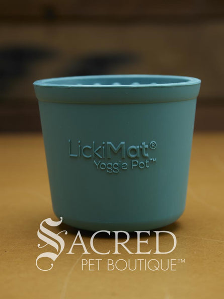 A teal silicone pet enrichment cup designed to hold food or treats for cats or dogs is sitting on a wooden table. The front of the cup is embossed with the brand name LickiMat and the product name Yoggie Pot. It has a smooth exterior and you are just able to see tge raised bumps of the interior. There is a white Sacred Pet Boutique watermark at the bottom of the image. 
