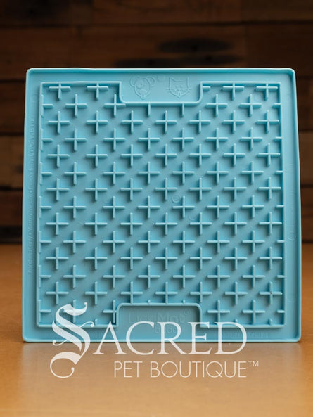 A blue LickiMat Boredom Buster designed for pets, with a textured surface designed to promote licking and calm behaviour. This design is the Classic Buddy and features a distinct criss cross matrix pattern designed to hold soft spreadable treats. It is on a wooden surface with a dark wood background and has a white Sacred Pet Boutique watermark.