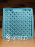 A blue LickiMat Boredom Buster designed for pets, with a textured surface designed to promote licking and calm behaviour. This design is the Classic Buddy and features a distinct criss cross matrix pattern designed to hold soft spreadable treats. It is on a wooden surface with a dark wood background and has a white Sacred Pet Boutique watermark.