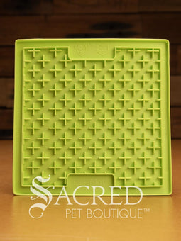 A purple LickiMat Boredom Buster designed for pets, with a textured surface designed to promote licking and calm behaviour. This design is the Classic Buddy and features a distinct criss cross matrix pattern designed to hold soft spreadable treats. It is on a wooden surface with a dark wood background and has a white Sacred Pet Boutique watermark.