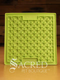 A green LickiMat Boredom Buster designed for pets, with a textured surface designed to promote licking and calm behaviour. This design is the Classic Buddy and features a distinct criss cross matrix pattern designed to hold soft spreadable treats. It is on a wooden surface with a dark wood background and has a white Sacred Pet Boutique watermark.