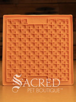 A purple LickiMat Boredom Buster designed for pets, with a textured surface designed to promote licking and calm behaviour. This design is the Classic Buddy and features a distinct criss cross matrix pattern designed to hold soft spreadable treats. It is on a wooden surface with a dark wood background and has a white Sacred Pet Boutique watermark.