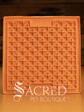 An orange LickiMat Boredom Buster designed for pets, with a textured surface designed to promote licking and calm behaviour. This design is the Classic Buddy and features a distinct criss cross matrix pattern designed to hold soft spreadable treats. It is on a wooden surface with a dark wood background and has a white Sacred Pet Boutique watermark.
