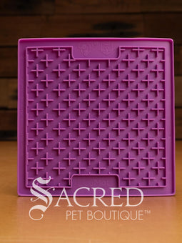 A purple LickiMat Boredom Buster designed for pets, with a textured surface designed to promote licking and calm behaviour. This design is the Classic Buddy and features a distinct criss cross matrix pattern designed to hold soft spreadable treats. It is on a wooden surface with a dark wood background and has a white Sacred Pet Boutique watermark.