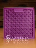 A purple LickiMat Boredom Buster designed for pets, with a textured surface designed to promote licking and calm behaviour. This design is the Classic Buddy and features a distinct criss cross matrix pattern designed to hold soft spreadable treats. It is on a wooden surface with a dark wood background and has a white Sacred Pet Boutique watermark.