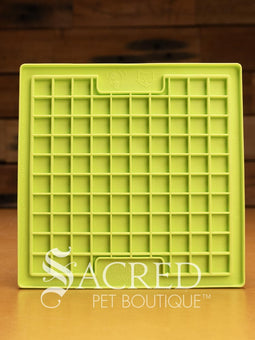 A purple LickiMat Boredom Buster designed for pets, with a textured surface designed to promote licking and calm behaviour. This design is the Classic Buddy and features a distinct criss cross matrix pattern designed to hold soft spreadable treats. It is on a wooden surface with a dark wood background and has a white Sacred Pet Boutique watermark.