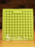 A green LickiMat Boredom Buster mat designed for pets, with a textured surface designed to promote licking and calm behaviour. This design is the Playdate and features a grid like pattern of small squares designed to hold soft spreadable treats. It is on a wooden surface with a dark wood background and has a white Sacred Pet Boutique watermark.
