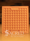 An orange LickiMat Boredom Buster mat designed for pets, with a textured surface designed to promote licking and calm behaviour. This design is the Playdate and features a grid like pattern of small squares designed to hold soft spreadable treats. It is on a wooden surface with a dark wood background and has a white Sacred Pet Boutique watermark.