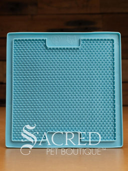 A purple LickiMat Boredom Buster designed for pets, with a textured surface designed to promote licking and calm behaviour. This design is the Classic Buddy and features a distinct criss cross matrix pattern designed to hold soft spreadable treats. It is on a wooden surface with a dark wood background and has a white Sacred Pet Boutique watermark.