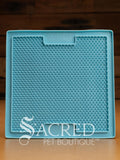 A blue LickiMat Boredom Buster mat designed for pets, with a textured surface designed to promote licking and calm behaviour. This design is the Sooter and features a surface that is covered in a dense grid of small, raised rubber bumps designed to trap soft food or treats to encourage licking. It is on a wooden surface with a dark wood background and has a white Sacred Pet Boutique watermark.