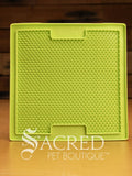 A green LickiMat Boredom Buster mat designed for pets, with a textured surface designed to promote licking and calm behaviour. This design is the Sooter and features a surface that is covered in a dense grid of small, raised rubber bumps designed to trap soft food or treats to encourage licking. It is on a wooden surface with a dark wood background and has a white Sacred Pet Boutique watermark.