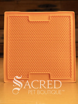 A purple LickiMat Boredom Buster designed for pets, with a textured surface designed to promote licking and calm behaviour. This design is the Classic Buddy and features a distinct criss cross matrix pattern designed to hold soft spreadable treats. It is on a wooden surface with a dark wood background and has a white Sacred Pet Boutique watermark.