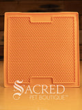 An orange LickiMat Boredom Buster mat designed for pets, with a textured surface designed to promote licking and calm behaviour. This design is the Sooter and features a surface that is covered in a dense grid of small, raised rubber bumps designed to trap soft food or treats to encourage licking. It is on a wooden surface with a dark wood background and has a white Sacred Pet Boutique watermark.