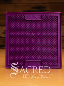A purple LickiMat Boredom Buster designed for pets, with a textured surface designed to promote licking and calm behaviour. This design is the Classic Buddy and features a distinct criss cross matrix pattern designed to hold soft spreadable treats. It is on a wooden surface with a dark wood background and has a white Sacred Pet Boutique watermark.