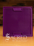 A purple LickiMat Boredom Buster mat designed for pets, with a textured surface designed to promote licking and calm behaviour. This design is the Sooter and features a surface that is covered in a dense grid of small, raised rubber bumps designed to trap soft food or treats to encourage licking. It is on a wooden surface with a dark wood background and has a white Sacred Pet Boutique watermark.