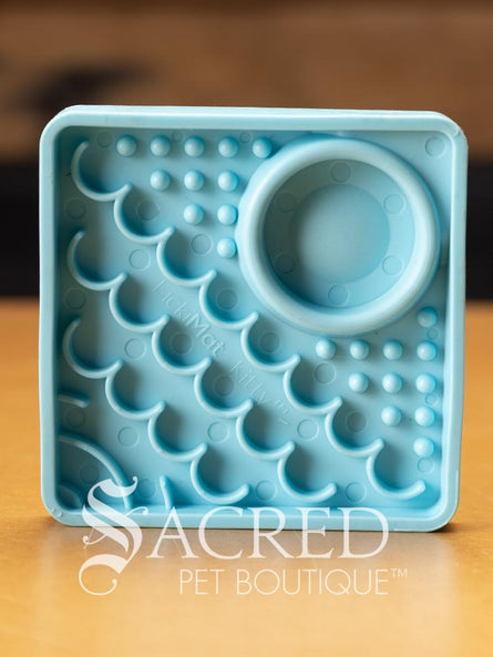 This blue LickiMat Kitty is a smaller version of the Catster and is an interactive slow-feeding enrichment mat designed for cats or kitttens. It features distinct functional sections. The right corner features small raised bumps with a circular micro-dish section for holding liquids and the left corner is covered in a series of layered, wave-like ridges. It is sitting on a wooden base with a dark wooden background. There is a white Sacred Pet Boutique watermark. 