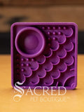 This purple LickiMat Kitty is a smaller version of the Catster and is an interactive slow-feeding enrichment mat designed for cats or kitttens. It features distinct functional sections. The left corner features small raised bumps with a circular micro-dish section for holding liquids and the right corner is covered in a series of layered, wave-like ridges. It is sitting on a wooden base with a dark wooden background. There is a white Sacred Pet Boutique watermark. 
