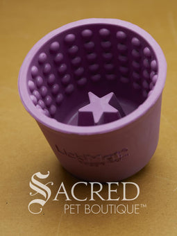 Four LickiMat Yoggie Pots of different colours are lined up on a wooden base with a wooden background. There is a teal, orange and purple one standing upright with a green one lying on top. There is a white Sacred Pet Boutique watermark at the bottom of the image. 
