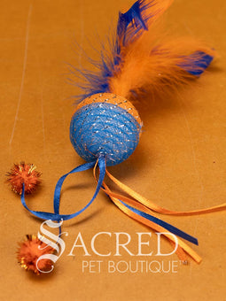 A selection of colourful mesh ball cat toys with ribbons and feather on a wooden background with a white Sacred Pet Boutique watermark. 