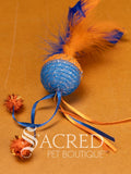 A single blue and orange mesh ball cat toy with ribbons and feathers on a wooden background with a white Sacred Pet Boutique watermark. 