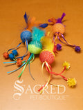 A selection of colourful mesh ball cat toys with ribbons and feather on a wooden background with a white Sacred Pet Boutique watermark. 