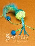 A single green and yellow mesh ball cat toy with ribbons and feathers on a wooden background with a white Sacred Pet Boutique watermark. 