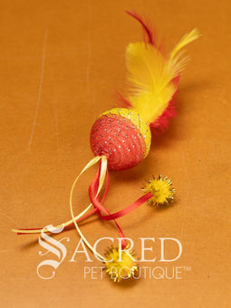 A selection of colourful mesh ball cat toys with ribbons and feather on a wooden background with a white Sacred Pet Boutique watermark. 